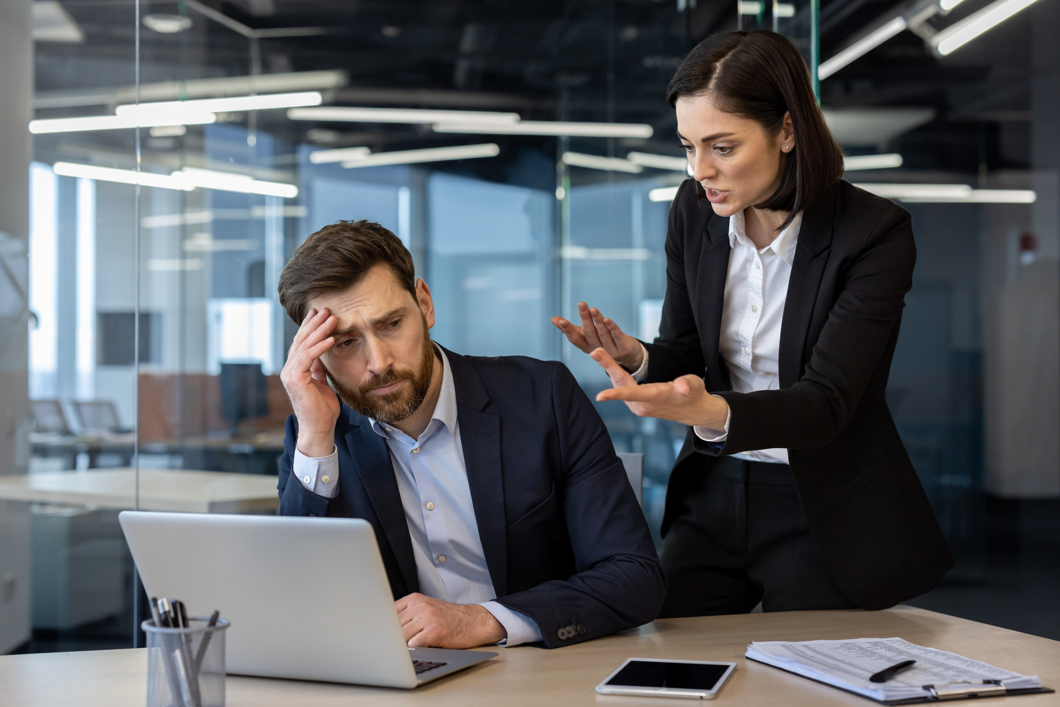 Conflicted office scene showing an angry female boss yelling at a frustrated male employee working on laptop. Tension highlights workplace stress and pressure during a business meeting