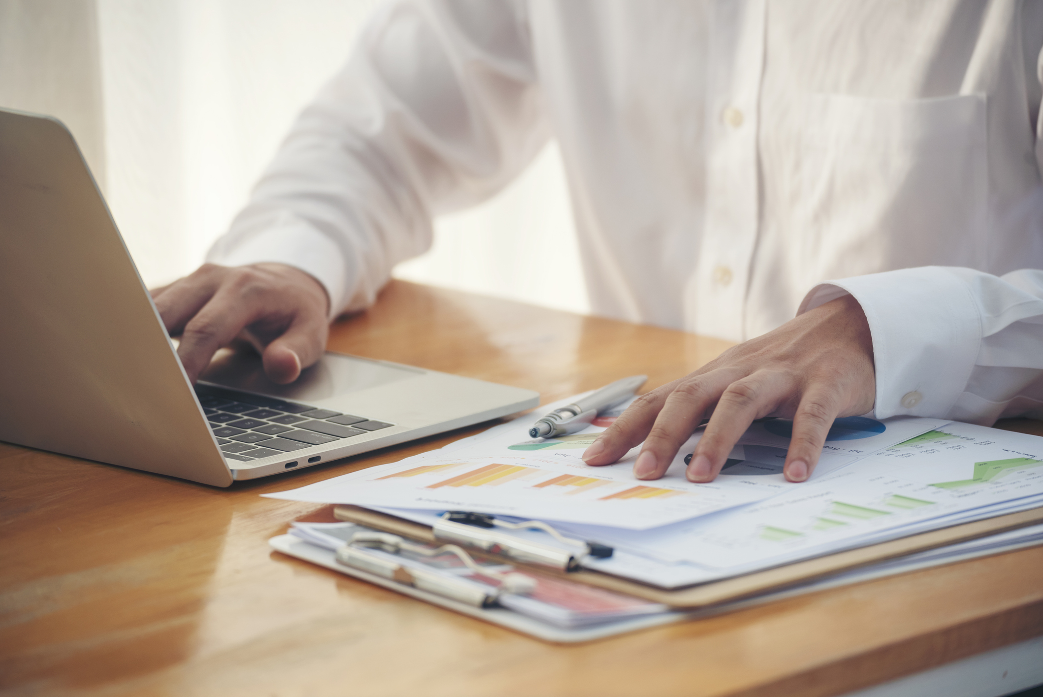Closeup businessman hands working on finance business document reading report paperwork spreadsheet using laptop at office desk. Man hands write on office note look at financial graph chart document